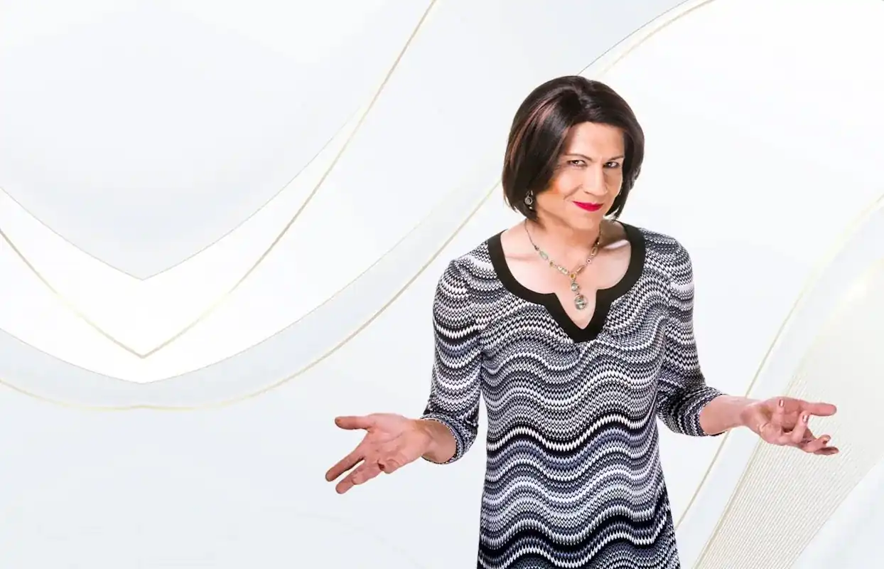 A confidently smiling cross dresser with short dark hair and a patterned dress, arms open in a welcoming gesture against a soft white and gold backdrop.